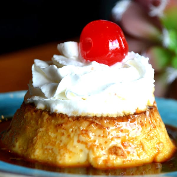 A Mexican flan dessert topped with whipped cream and a bright red cherry on a blue plate.