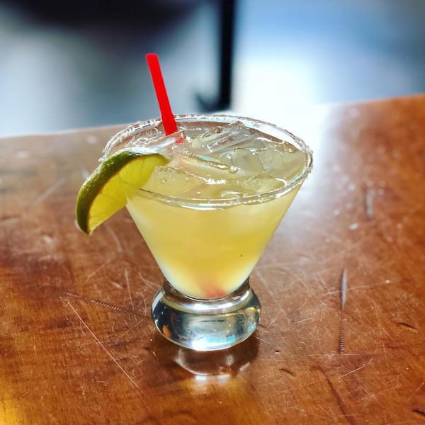 A margarita with a salted rim, lime wedge, and red straw, on a wooden table.